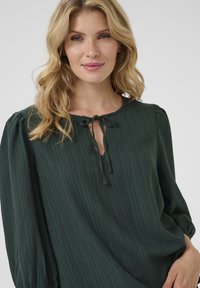 Blusa verde scuro realizzata in tessuto testurizzato, con scollatura rotonda, dettaglio con fiocco e maniche a palloncino con polsini elastici.