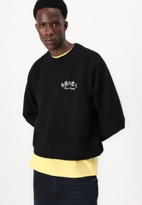Sort sweatshirt med oversized pasform, prydet med hvidt logo og japansk tekst på brystet, lagdelt over en gul t-shirt, parret med mørke jeans.