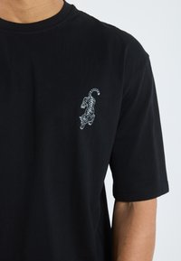 T-shirt noir en coton, avec un design de tigre brodé en blanc sur la partie gauche de la poitrine. Manches courtes et coupe standard.