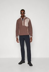 Brun fleece-genser med kvartglidelås og beige detaljer, kombinert med mørke bukser. Modellen har på seg solbriller og svarte sko. Jevn tekstur.