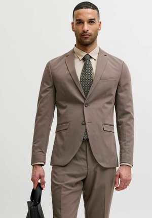 Man in taupekleurig pak, met een patroon das en beige overhemd, die een zwarte, gestructureerde handtas vasthoudt, staand tegen een effen lichte achtergrond.