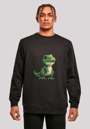 Junger Mann trägt einen schwarzen Pullover mit einem grünen Cartoon-T-Rex-Dinosaurier-Motiv in der Mitte, steht vor einem hellgrauen Hintergrund.