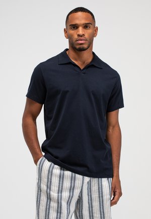 Man draagt een marineblauw poloshirt met korte mouwen en witte broek met verticale blauwe strepen, staand met één hand in de zak.