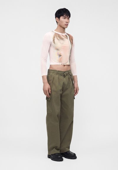 Νέος άντρας που φοράει διάφανο crop top με μακριά μανίκια και αφηρημένο σχέδιο προσώπου, χακί cargo παντελόνια και μαύρα chunky παπούτσια, στέκεται όρθιος.