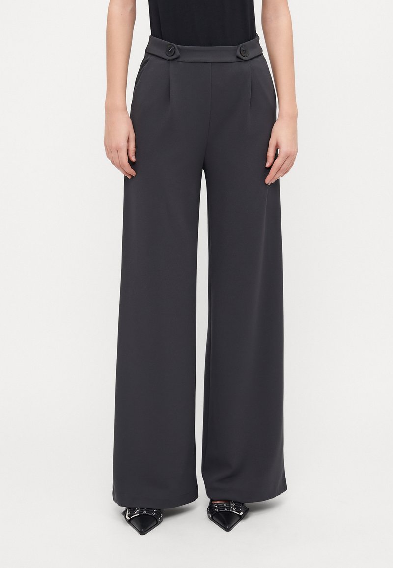 Femme portant un pantalon gris foncé taille haute à jambes larges avec des boutons à la ceinture et des chaussures noires pointues avec des boucles argentées.