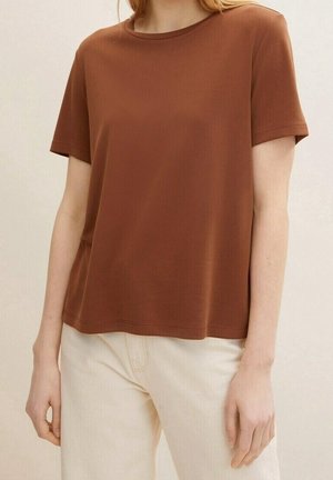 Kvinde iført en brun T-shirt med korte ærmer og lyse beige bukser, stående foran en ensfarvet lys baggrund.