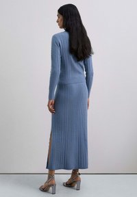 Mujer con cabello largo y oscuro, vistiendo un suéter de punto en color azul claro y una falda larga a juego con una abertura lateral, de pie contra una pared clara y lisa.