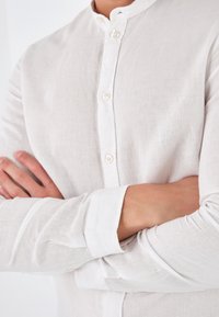 Camicia bianca in lino con colletto alla coreana, bottoni sul davanti e maniche lunghe. La trama è leggera e leggermente stropicciata.