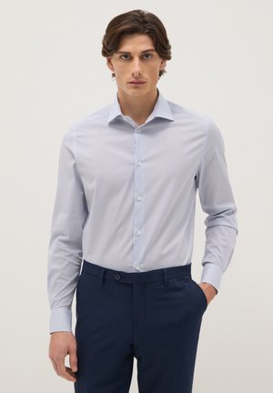 Junger Mann trägt ein hellblau gestreiftes Hemd, das in marineblaue Anzughose gesteckt ist, steht mit einer Hand in der Tasche.