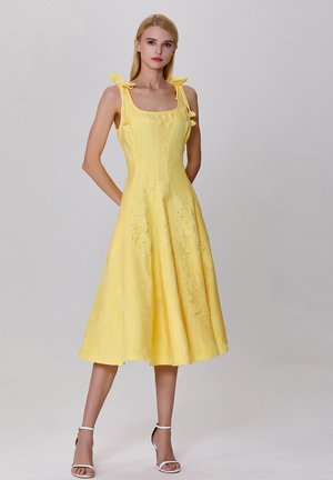 Robe midi jaune avec un décolleté carré, des bretelles à nouer et une jupe évasée. Le tissu a une texture subtile et un doux éclat, idéal pour les temps chauds.