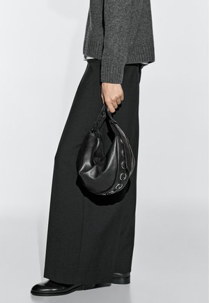 Person in schwarzen Hosen mit weitem Bein und grauem Pullover, die eine kleine schwarze Lederhandtasche mit silbernen Ringakzenten hält.