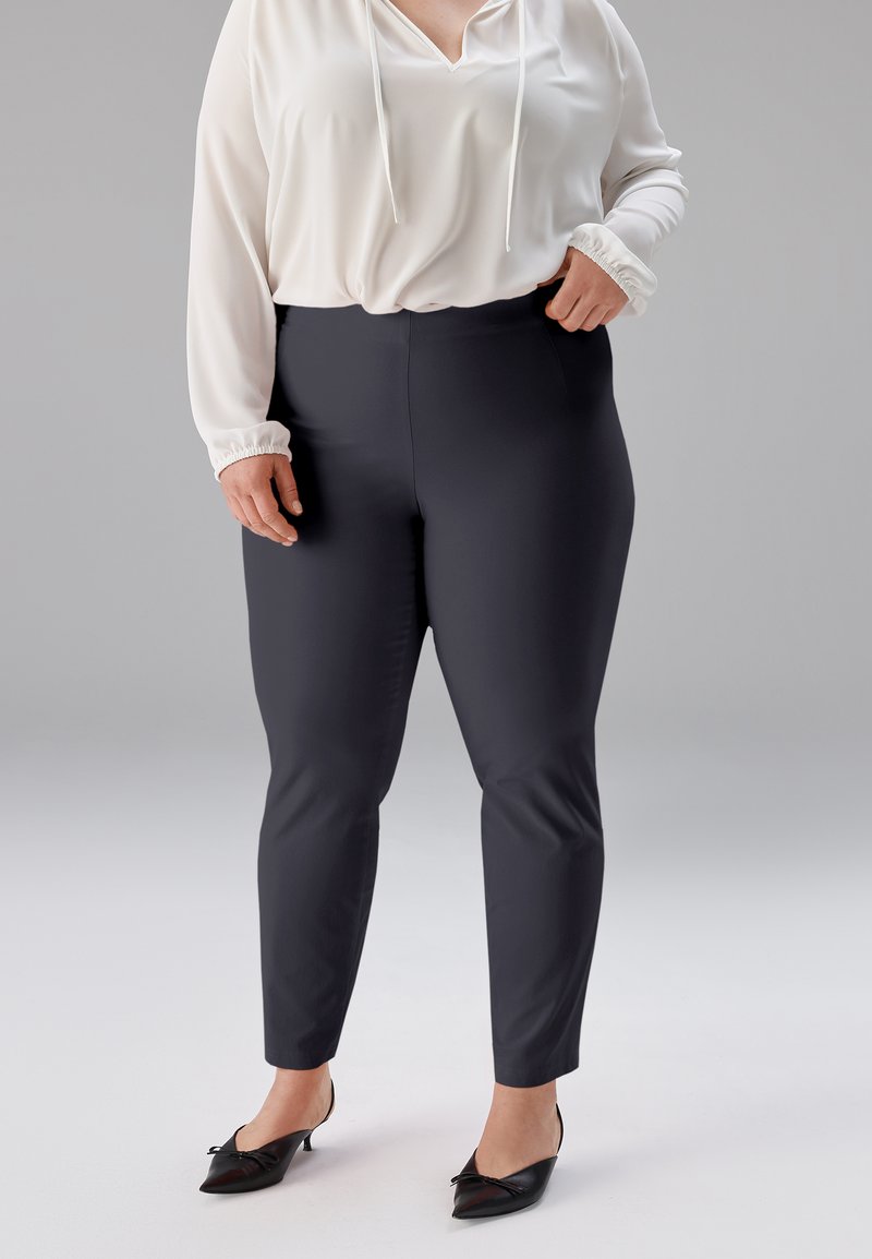 Schwarze, eng anliegende Hose mit glatter Textur und hohem Bund, kombiniert mit einer weißen Bluse mit V-Ausschnitt und Kordelzug.