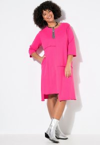 Abito fucsia con maniche a 3/4, dettagli con tasche e orlo irregolare, abbinato a stivali alla caviglia metallici argento. Tessuto dalla consistenza liscia.