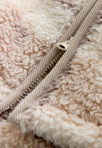 Puszysty beżowy materiał z teksturowanymi pętlami, z kremowym zamkiem błyskawicznym i prążkowanym wykończeniem wzdłuż krawędzi. Miękka, ciepła i przyjemna w dotyku powierzchnia.