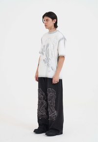 Přepychové bílé tričko s grafickým motivem šedého orla a textem, spárované s černými kalhotami s širokými nohavicemi, které mají složité vyšité vzory.