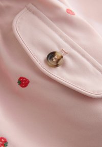 Pink fabric with fine texture featuring an embroidered red strawberry design and a glossy brown button on a side pocket.