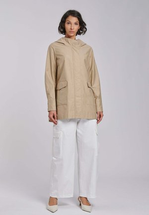 Kobieta o kręconych, ciemnych włosach, ubrana w beżową kurtkę z kapturem, białe szerokie spodnie typu cargo i białe szpiczaste szpilki, stojąca na jednolitym tle.