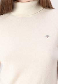 Krémový rolákový svetr z měkké pleteniny s žebrovaným límcem. Obsahuje malé logo na levé straně, minimalistický design.