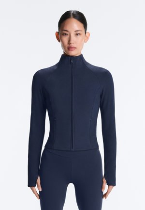 Frau mit dunklen Haaren, die eine enganliegende marineblaue Jacke mit Reißverschluss und passende Leggings vor einem schlichten hellen Hintergrund trägt.