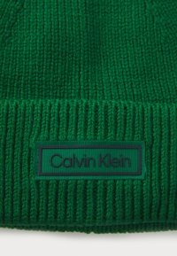 Zelená pletená čiapka s rebrovanou textúrou a plochou, obdĺžnikovou nášivkou loga Calvin Klein v tmavozelenej farbe na leme.