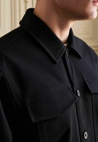 Sort bomuldsskjortejakke med spids krave, knaplukning og to brystlommer. Har en glat tekstur og robust konstruktion.