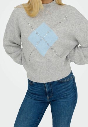 Kvinde iført grå strikket sweater med blåt diamantmønster og rhinsten, kombineret med blå jeans, stående med hænderne på hofterne.