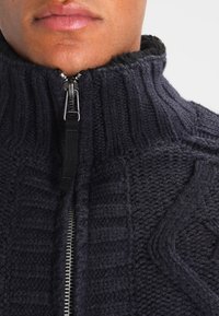 Námořnický pletený svetr na zip s ribovaným límcem, texturovanými vzory a stříbrn�ým zipem. Měkká příze a vysoký límec.