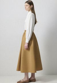 Camisa blanca de manga larga con hombros fruncidos, combinada con una larga falda mostaza en forma de A. Unos zapatos planos de tejido en marrón claro completan el conjunto.