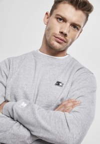 Sudadera gris con cuello redondo y puños acanalados. Presenta un pequeño logo negro en el pecho y una tela suave y texturizada.