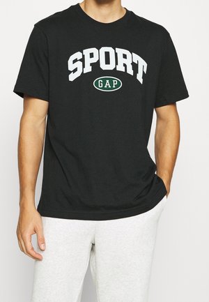 Mężczyzna nosi czarną koszulkę z krótkim rękawem z białym napisem „SPORT” oraz zielonym owalnym logo „GAP”, w połączeniu z jasnoszarymi spodniami.