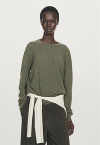 Olivgrön stickad tröja med bred halsringning, ihopparad med mörkbruna byxor som har ett vitt knutet bälte knutet i midjan för detaljer.