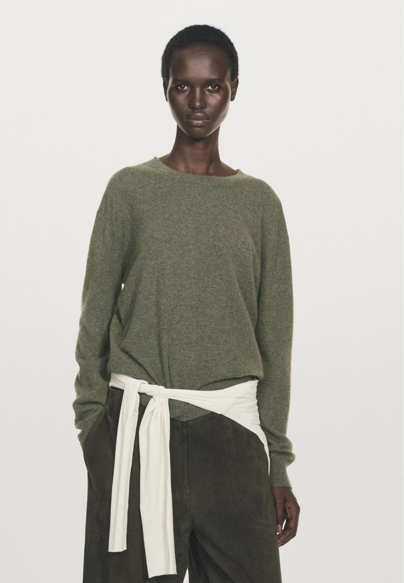 Olivgrön stickad tröja med bred halsringning, ihopparad med mörkbruna byxor som har ett vitt knutet bälte knutet i midjan för detaljer.