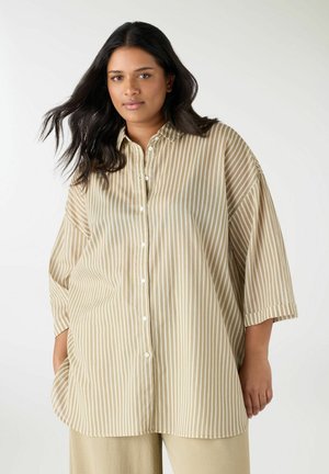 Donna con lunghi capelli scuri che indossa una camicia beige chiaro a righe verticali bianche e beige morbida con bottoni e pantaloni beige chiaro, in piedi davanti a uno sfondo bianco.