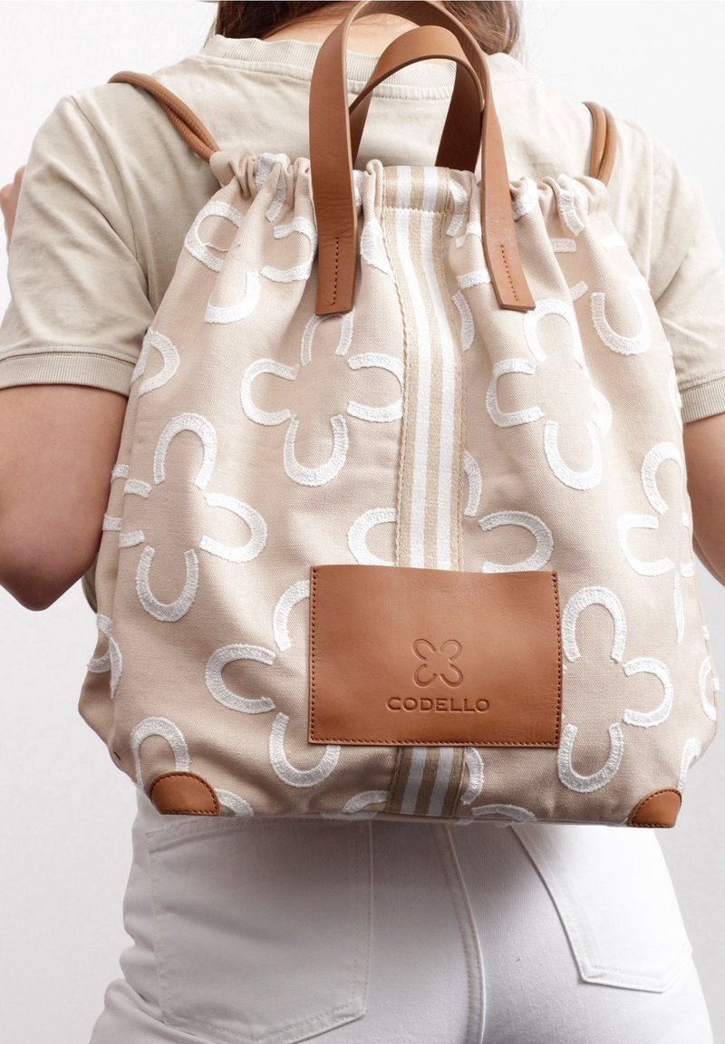 Zaino beige con motivo floreale bianco e dettagli in pelle marrone chiaro. Presenta una chiusura a coulisse e una toppa con logo sul davanti.