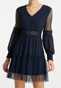 Robe bleu marine avec un col en V et des manches en mesh. Dotée d'une taille en dentelle et d'un ourlet volanté en tulle. S'étend doucement sous le genou.