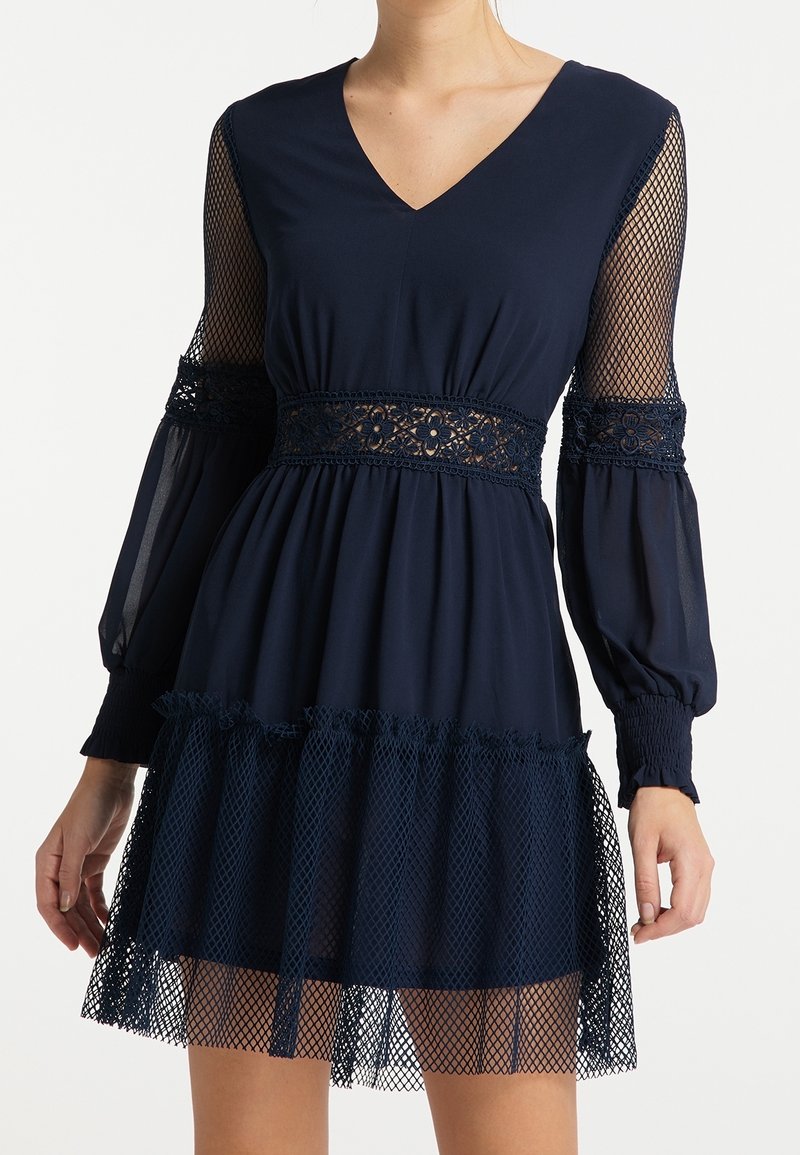 Robe bleu marine avec un col en V et des manches en mesh. Dotée d'une taille en dentelle et d'un ourlet volanté en tulle. S'étend doucement sous le genou.