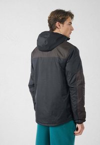 Jonge man die een zwart-bruine capuchonjas draagt, gezien van achteren, gecombineerd met teal gekleurde broek tegen een effen lichte achtergrond.