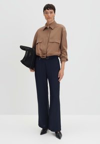 Bruine blouse met kraag en aantrekkoord in de taille, twee voorzakken, gecombineerd met marineblauwe wijde broekspijpen en zwarte puntige hakken. Houdt een zwarte mesh tas vast.