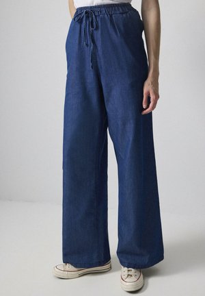 Wide-leg jeans in dark blue denim with an elastic waistband and drawstring, featuring a smooth texture and a relaxed fit. Paired with beige sneakers.