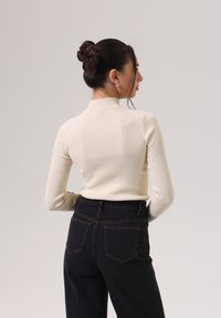 Rippstrick-Creme-Turtleneck und hochtaillierte schwarze Jeans mit kontrastierenden orangefarbenen Nähten; Figur von hinten gezeigt, mit hochgestecktem Haar.