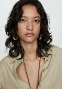 Beige button-up shirt with a cutout detail at the collar, worn with silver hoop earrings. Dark, wavy hair frames the face. Neutral background.