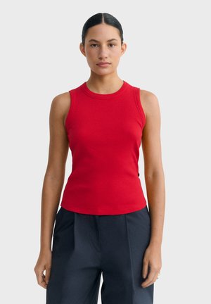 Jeune femme aux cheveux foncés tirés en arrière, portant un haut sans manches rouge côtelé et un pantalon noir ample, debout devant un fond uni.