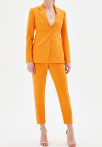 Pomarańczowy komplet garniturowy z dopasowaną marynarką i wąskimi spodniami; gładka faktura, zapięcie na dwa guziki oraz subtelne poduszki na ramionach.