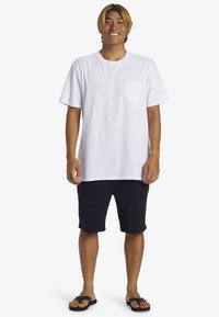 Camiseta blanca de algodón con mangas cortas y un bolsillo en el pecho, combinada con pantalones cortos azul marino y chanclas negras, de pie sobre un fondo liso.