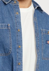 Camicia in denim blu con bottoni dorati, due tasche sul petto e un piccolo patch con logo. Il tessuto ha una texture liscia con sottili dettagli di cucitura.