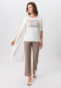 Witte top met decoratief beige patroon, gecombineerd met beige broeken met splitten aan de onderkant en witte sandaaltjes met bandjes. Vloeiende witte buitenlaag.