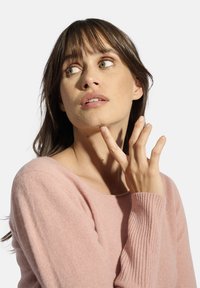 Světle růžový, žebrovaný svetr s širokým výstřihem. Žena, která gestikuluje poblíž brady, má dlouhé vlasy a decentní make-up, na jednobodovém pozadí.