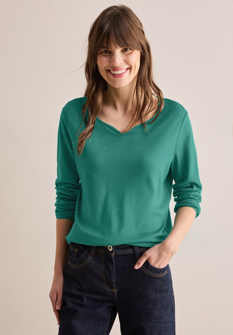 Vihreä pitkähihainen paita, jossa on V-pääntie, pehmeä tekstuuri ja kääntöhiharesorit. Yhdistetty tummiin farkkuihin; kädet taskuissa. Neutraali tausta.