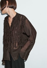 Donna con capelli corti castani, che indossa una blusa trasparente marrone scuro con rouches verticali texturizzate e pantaloni neri, voltata parzialmente di lato.