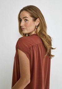 Blusa color ruggine con maniche corte, schiena drappeggiata e texture liscia. La modella si gira leggermente, mostrando capelli morbidi e orecchini a cerchio argentati.